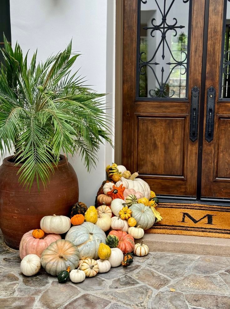Front door fall display with heirloom pumpkins and gourds beside a palm plant and ornate wooden doors